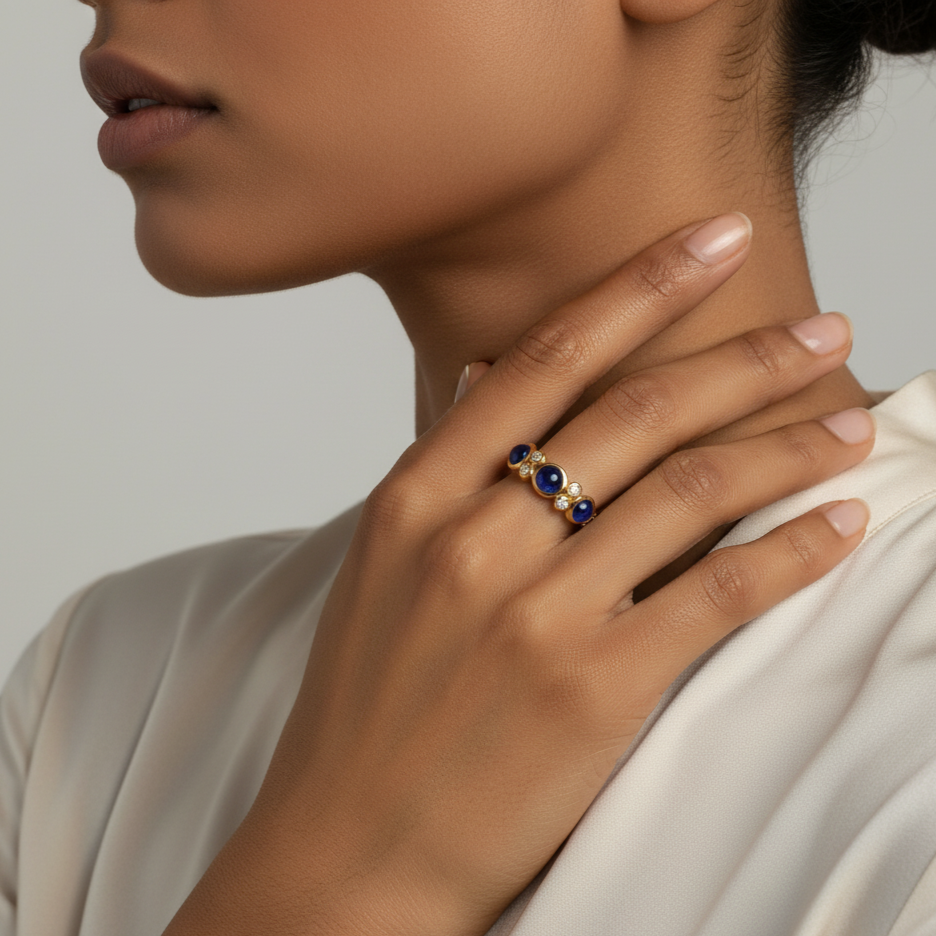 Close-up of a hand wearing a gold ring with blue stones, touching the neck against a neutral background.