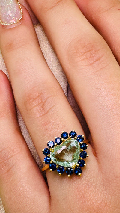Paraiba Tourmaline Heart shape And Blue Sapphire Round Ring In 18K Yellow Gold - Tresor Collection Close-up of Paraiba tourmaline heart ring with sapphire halo worn on hand – glowing neon-blue gemstone