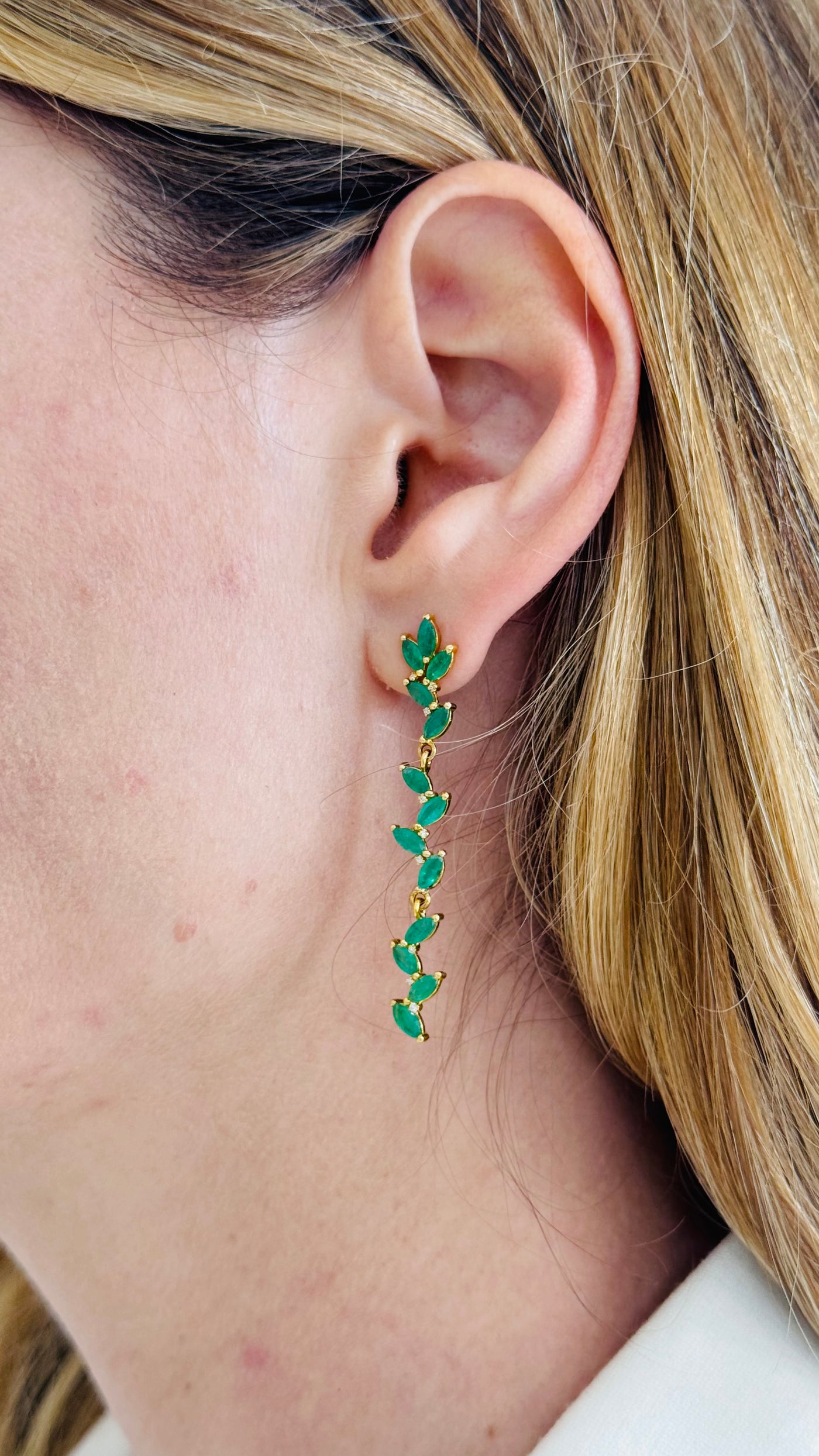 Close-up of a person wearing green earrings with gold accents. Side view of model wearing long emerald drop earrings – elegant fine jewelry look