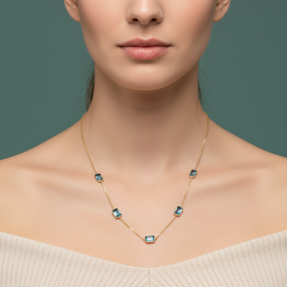Necklace with blue gemstones worn by a model on a plain background Close-up of Blue Topaz station necklace on model’s neck – luxury fine jewelry styling