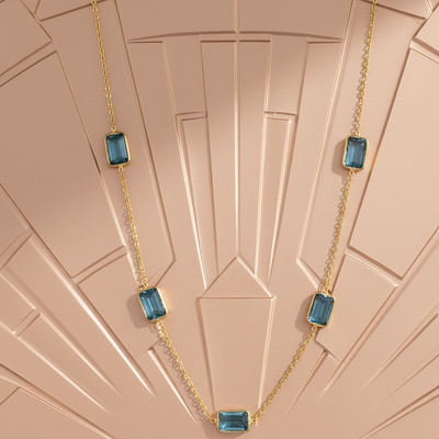 Gold necklace with blue gemstones on a patterned pink background Blue Topaz station necklace styled on silk fabric – premium handcrafted jewelry