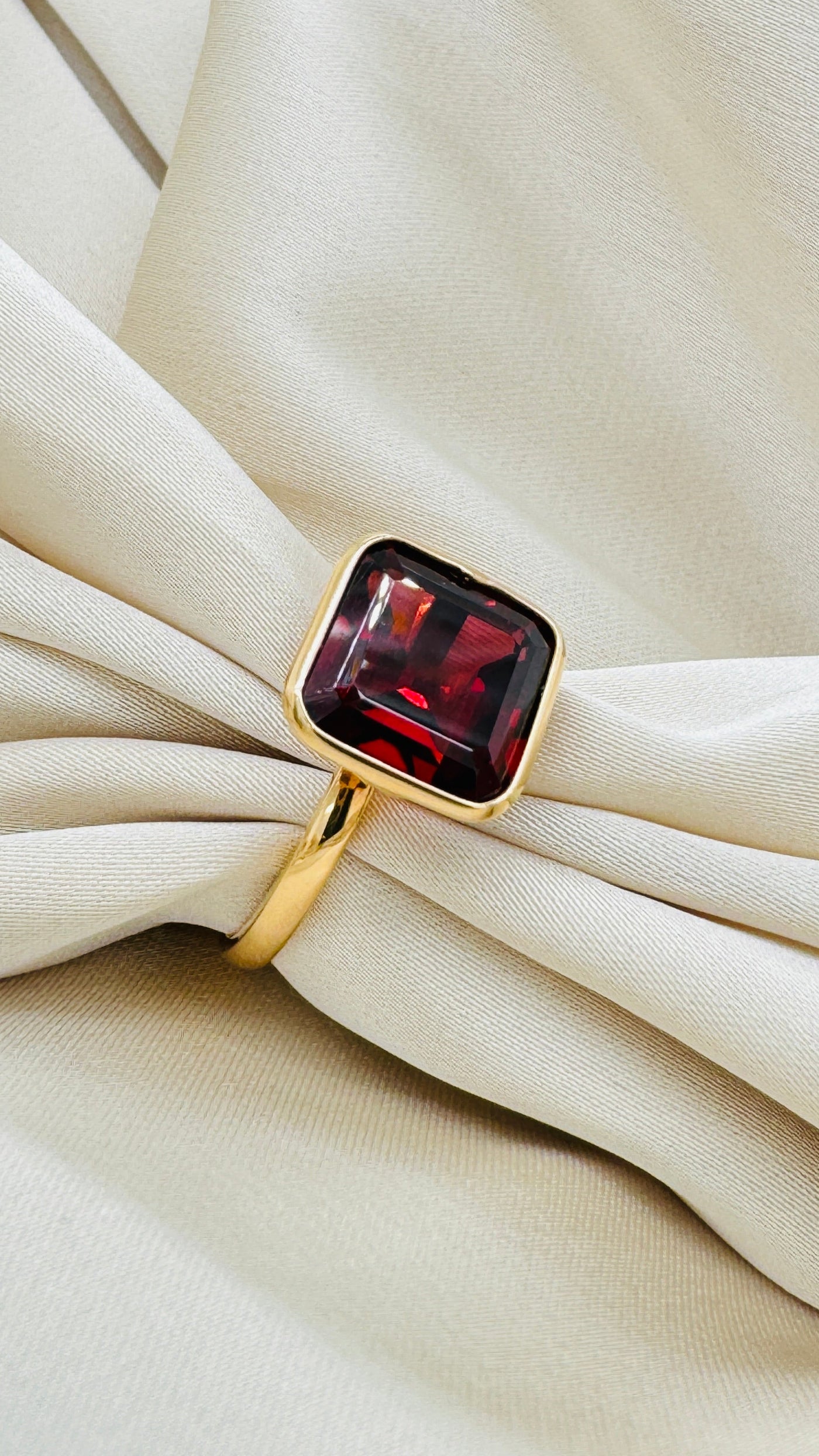 Gold ring with red gemstone on beige fabric background Artistic angle shot of 18K gold rhodolite garnet ring – elegant handcrafted gemstone jewelry by Tresor Collection