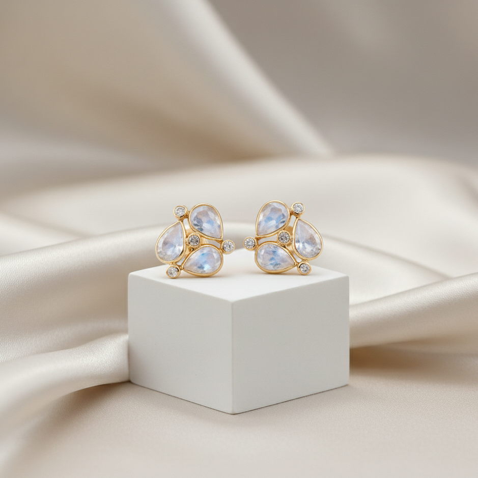 Pair of gold earrings with clear gemstones on a white cube against a beige fabric background Rainbow Moonstone stud earrings in 18K gold displayed on jewelry box – timeless gemstone elegance