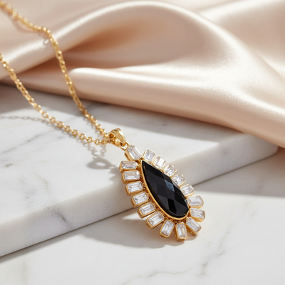 Gold necklace with a black teardrop pendant on a marble surface Black gemstone teardrop pendant with gold chain and pearl accents – luxury jewelry flatlay by Tresor Collection
