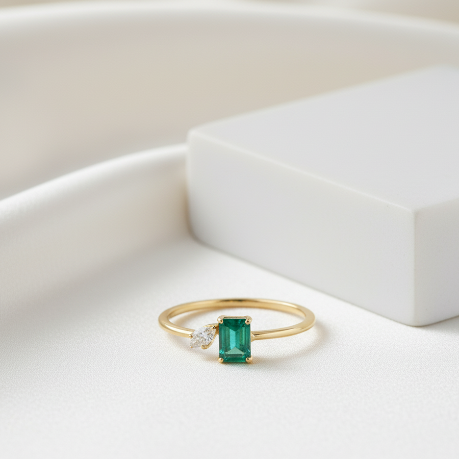 Gold ring with a green gemstone on a white surface Emerald and pear-diamond gold ring displayed on jewelry tray – modern fine-jewelry styling