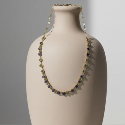 Necklace draped over a beige ceramic vase on a gradient 18K gold Tanzanite & Tsavorite necklace displayed on stand – handcrafted gemstone jewelry by Tresor Collectionbackground