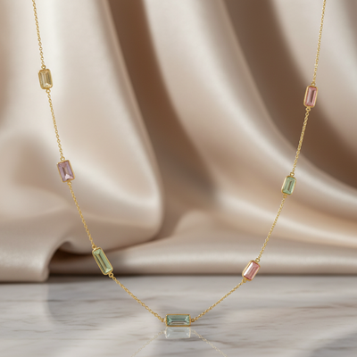 Gold necklace with green and pink gemstones on a beige fabric background Close-up of 18K gold multicolor baguette necklace – handcrafted fine jewelry from Tresor Collection boutique