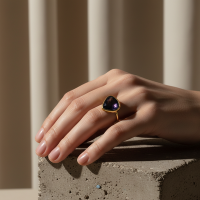 Hand wearing a ring with a purple gemstone on a neutral background Elegant blue sapphire ring on model – designer gold jewelry for women by Tresor Collection