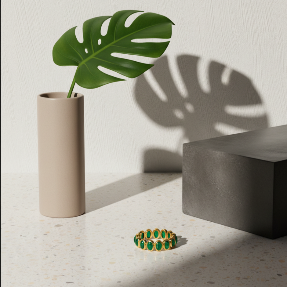 Green ring on a light surface with a beige vase and plant leaf casting shadows. Close-up of 18K gold oval cabochon emerald ring – handcrafted fine jewelry from Tresor Collection boutique